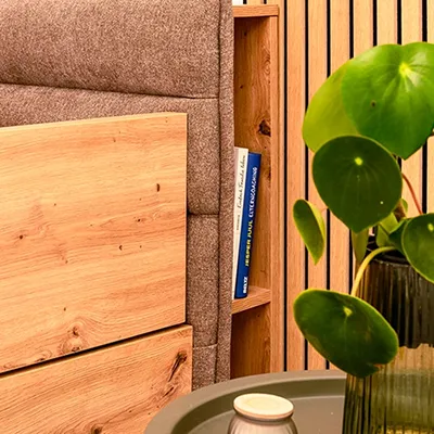 Detailgetreue Interieur-Fotografie: Familienbett-Ecke. Perfekte Inszenierung für Schlafzimmerkollektionen und Wohnmagazine. Detailaufnahme eines Familienbettes: Holzrahmen, Stoffkopfteil, Beistelltisch mit Pflanze und Büchern. Authentische Schlafzimmer-Interieur-Fotografie.